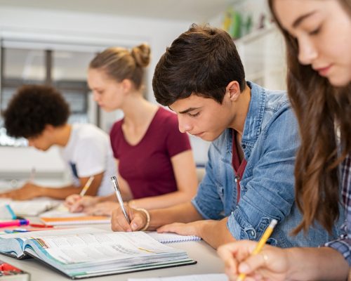 Group of college students studying in classroom writing notes during lesson. Focused guy and girls studying in college library sitting at desk. Group of multiethnic university students doing research sitting in a row.